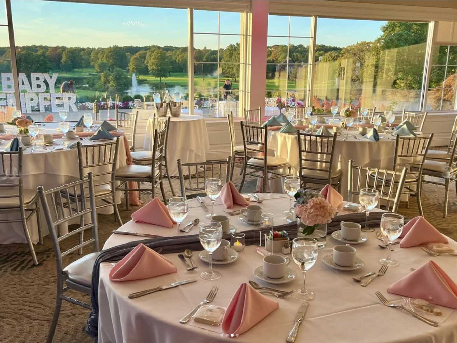 baby-shower-reception Elegant event space perfect for NJ Milestone events, featuring round tables set with pink napkins, glassware, and floral centerpieces. Large windows offer a view of the green landscape. Silver chairs elegantly surround the tables, completing the sophisticated ambiance.
