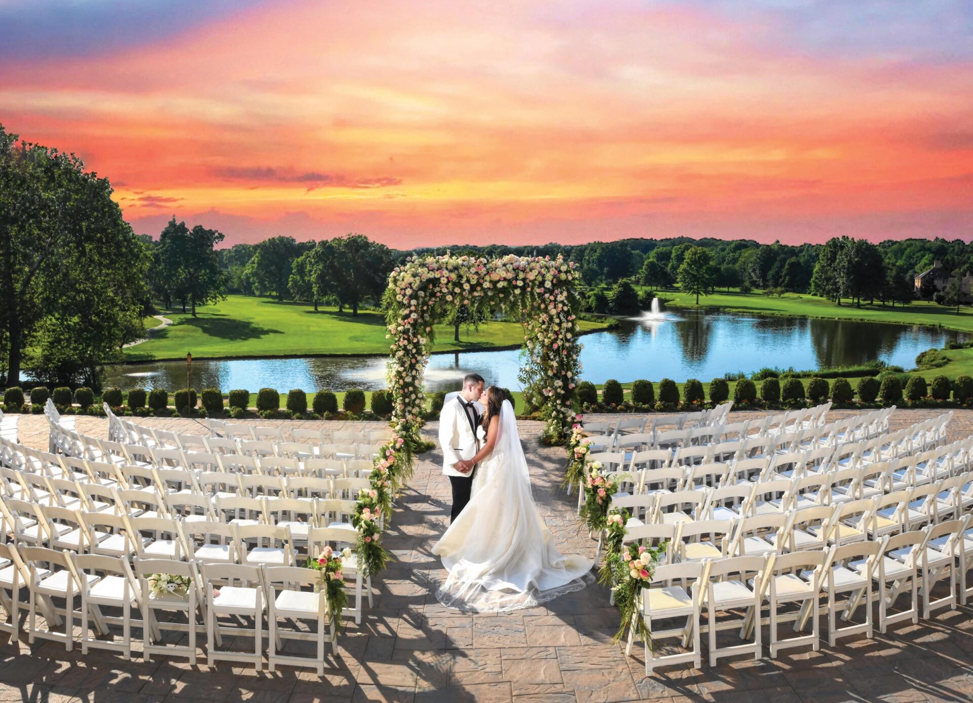 Brooklake-sunset-wedding-ceremony Bride and groom kiss under a floral arch at a stunning NJ wedding venue. Rows of white chairs line the pathway, while a pond and sunset sky provide the perfect backdrop.