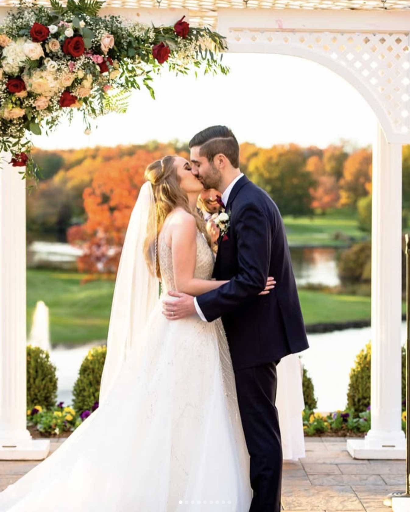 autumn-wedding-ceremony-at-brooklake A bride and groom kiss under a floral arch at an outdoor NJ wedding & event venue, with a lake and vibrant autumn trees in the background.