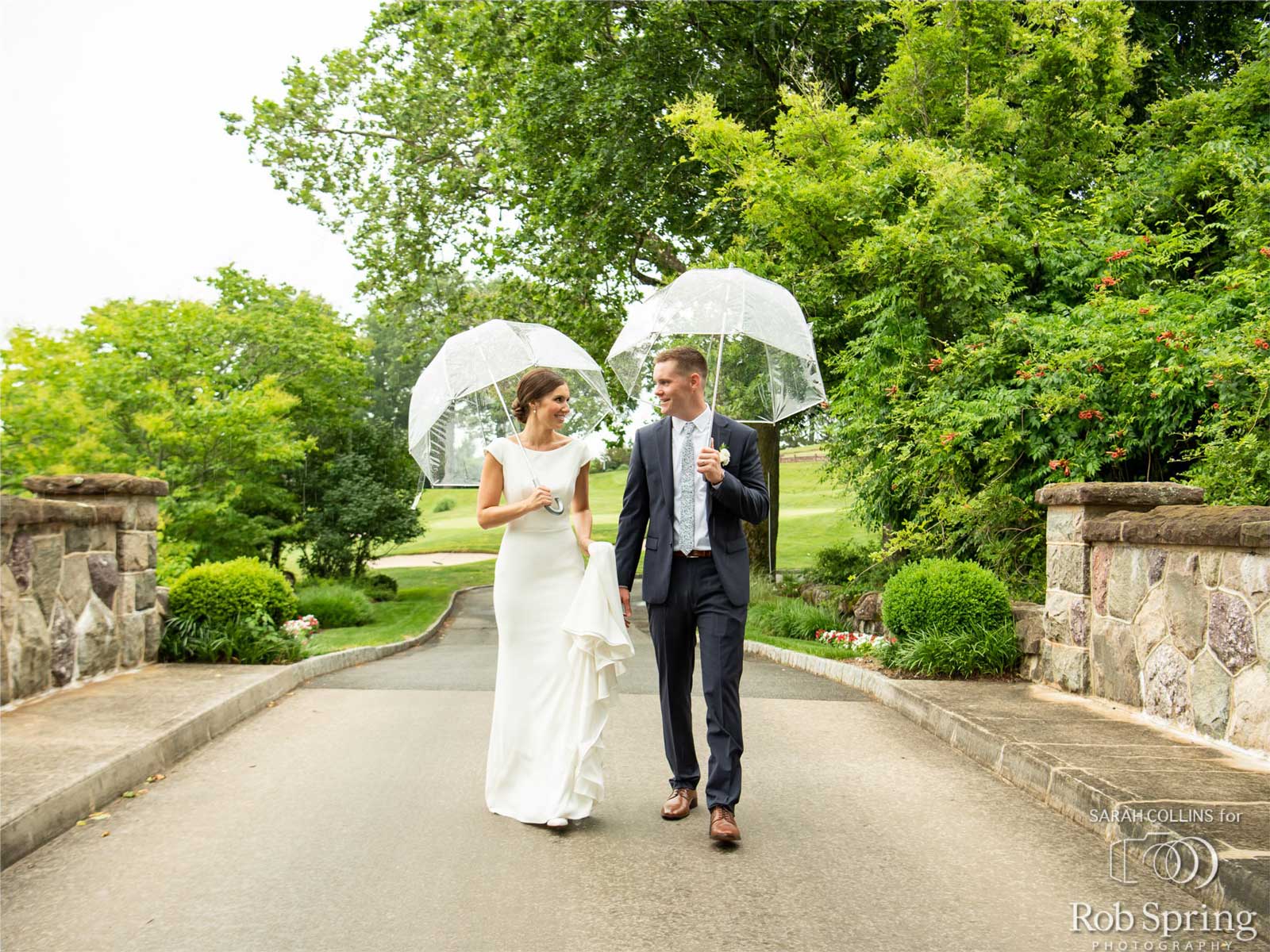 A bride and groom walk together on a paved path at an NJ wedding venue, holding clear umbrellas, surrounded by greenery and stone walls on a cloudy day.