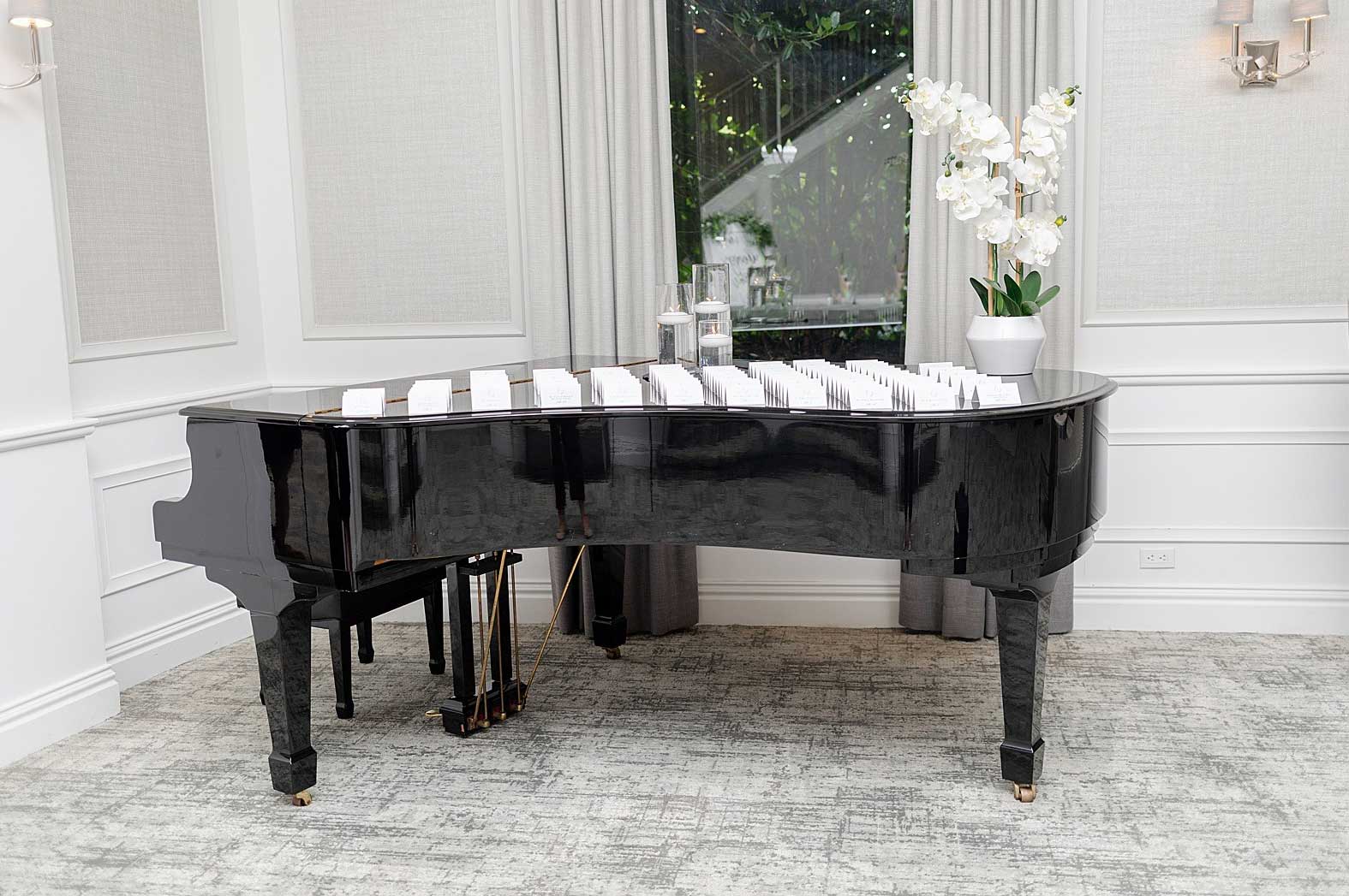 brooklake-foyer-piano-place-cards A black grand piano at this NJ wedding venue displays rows of white place cards, with two candles and a white orchid arrangement on top, in a bright room with large windows.