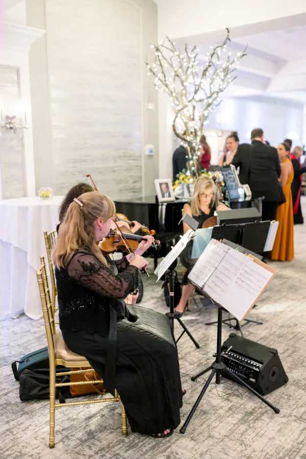 brooklake-musicians-cocktail-hour A small classical music ensemble performs indoors at an nj wedding venue, with a violinist, cellist, and pianist playing near a decorated tree as guests mingle in the background.