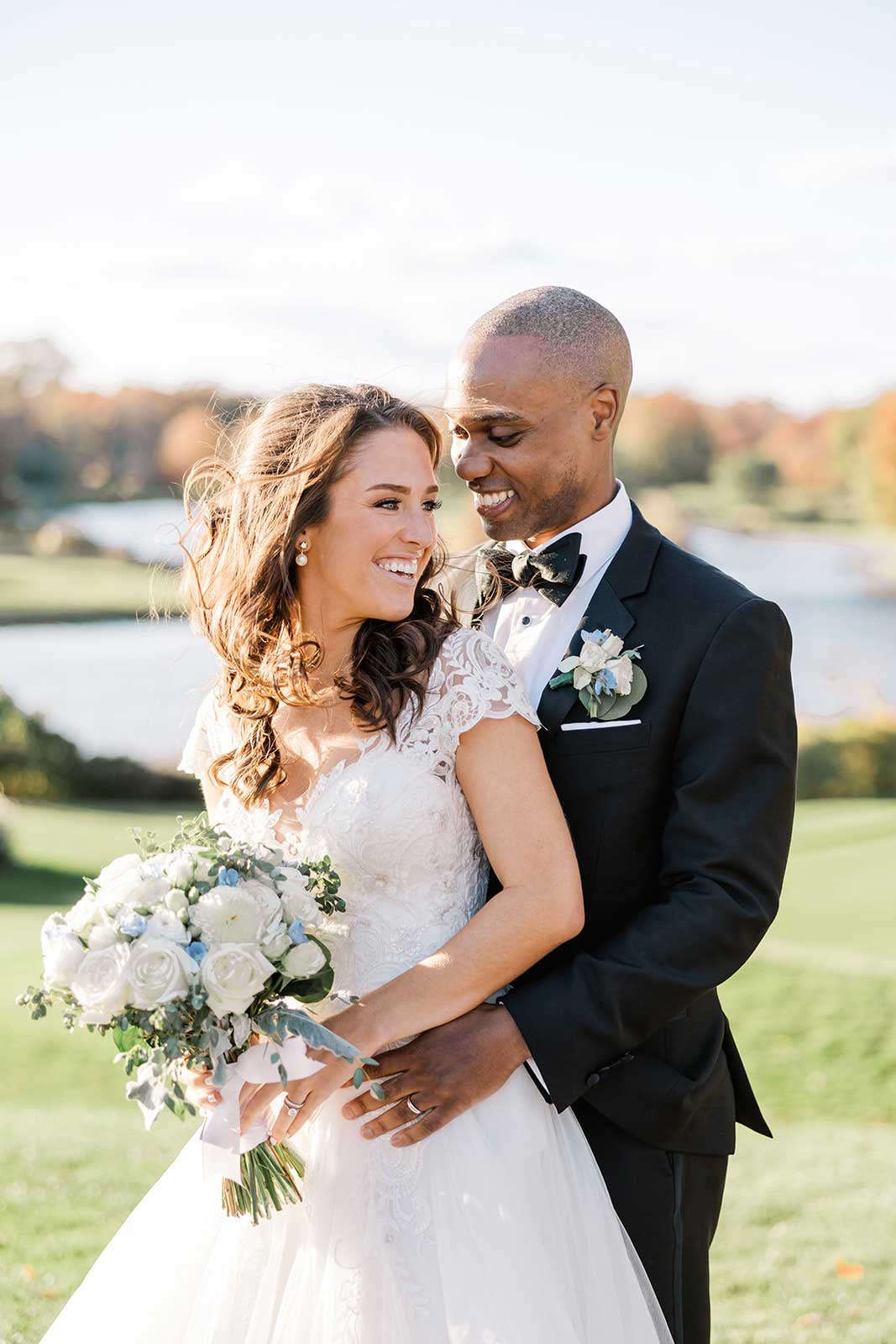 A bride in a white dress holding a bouquet stands with her groom in a tuxedo, smiling together outdoors by a pond at an elegant NJ wedding & event venue.