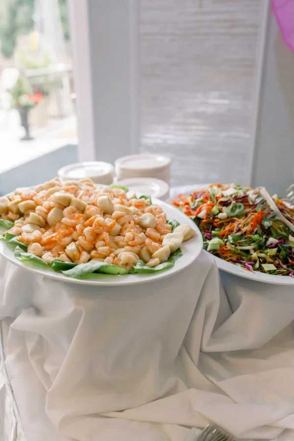 brooklake-seafood-cocktail-hour A platter of shrimp on lettuce sits next to a bowl of colorful salad with shredded vegetables, both beautifully arranged on a white tablecloth—perfect inspiration for your NJ wedding venue catering.