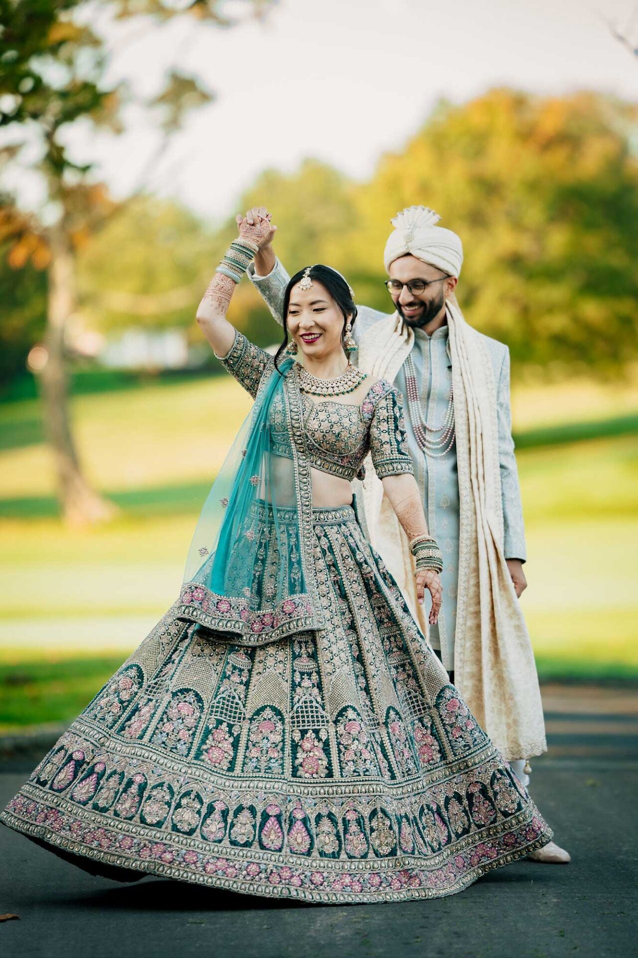 A bride in a detailed lehenga and a groom in a sherwani and turban pose outdoors at an elegant NJ wedding & event venue, with the groom spinning the bride on a paved path.