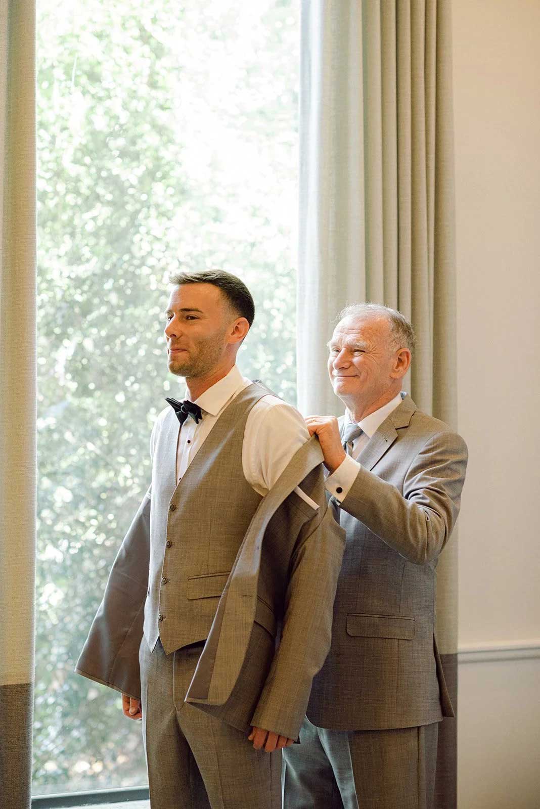brooklake-upasanamainali-groom-getting-ready An older man helps a younger man put on a suit jacket in a brightly lit room at an elegant NJ wedding venue, both dressed formally in grey suits.