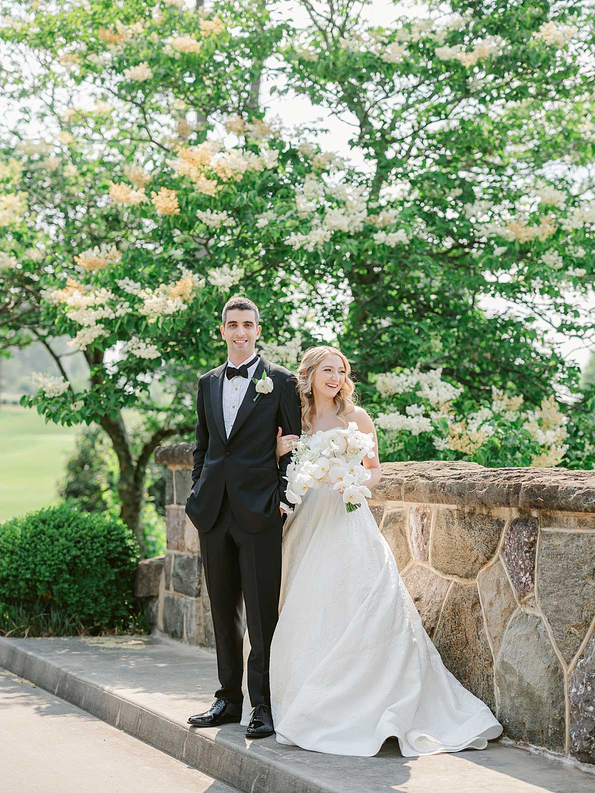 A bride and groom stand side by side outdoors near a stone wall at an elegant NJ wedding & event venue, dressed in formal wedding attire, with greenery and blooming trees in the background.