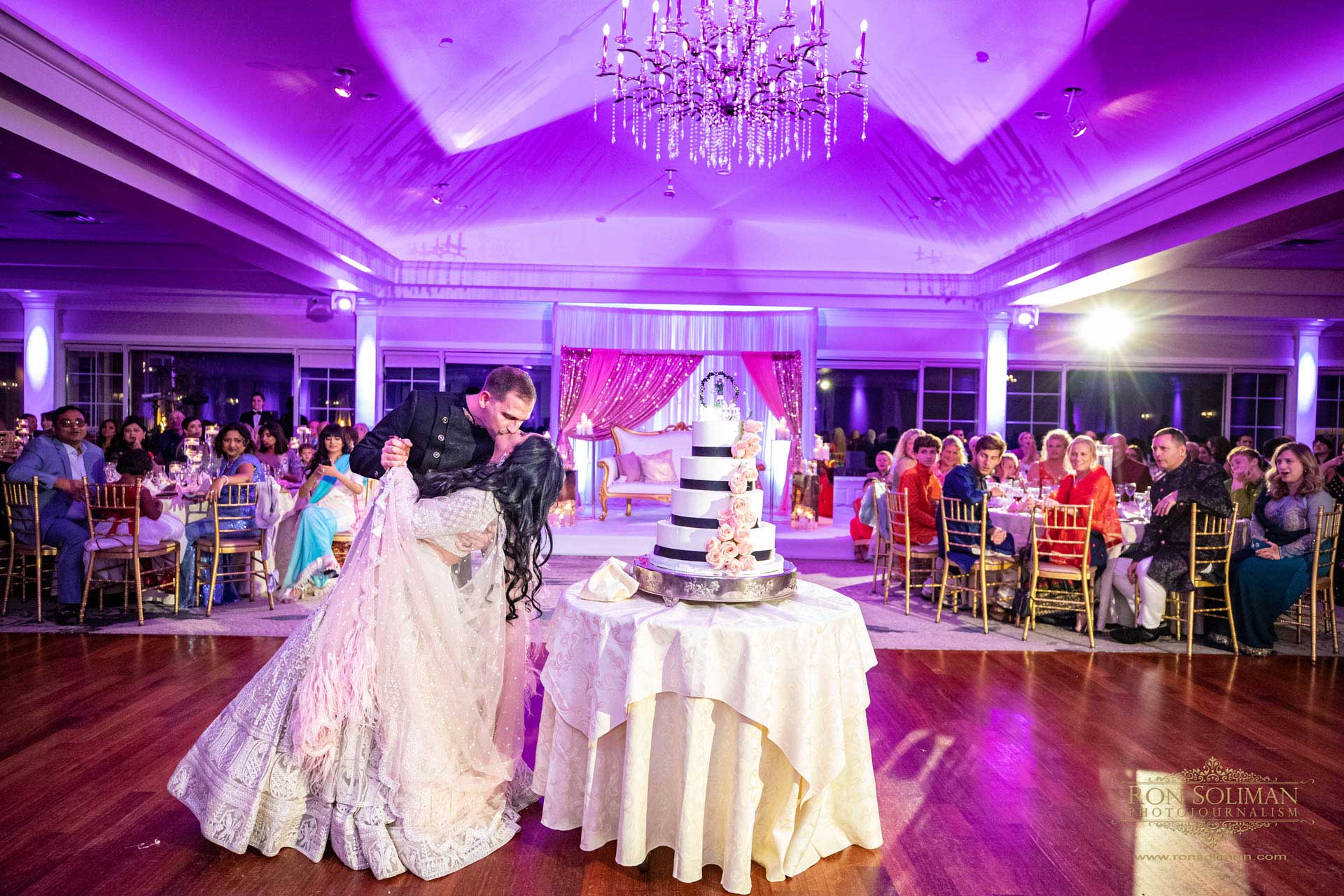 A couple shares a dip on the dance floor at a wedding reception, with guests seated around and a multi-tiered cake in the foreground—capturing a magical moment reminiscent of joyful NJ Teen Events.