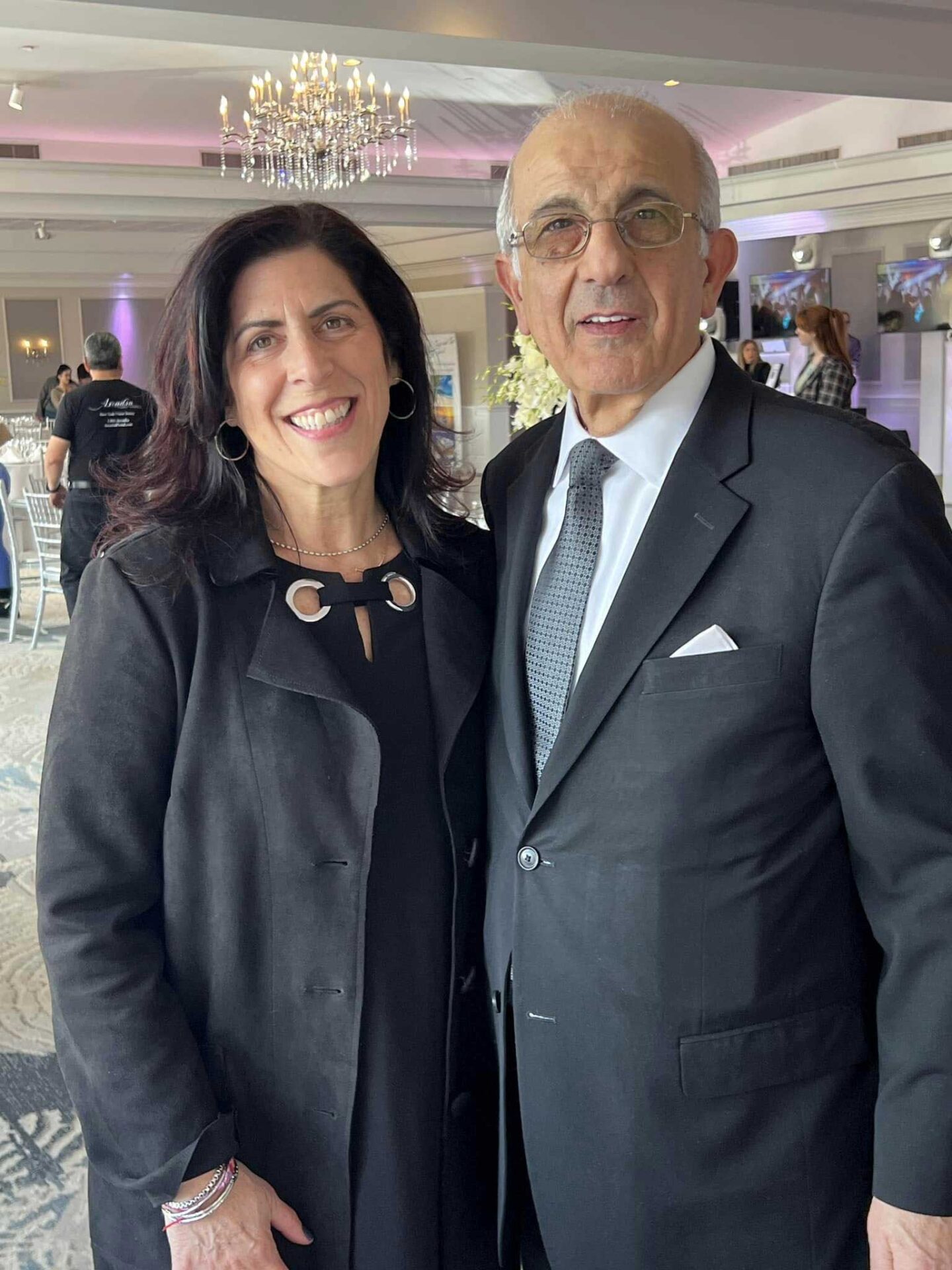 A woman and a man in formal attire pose together and smile at an indoor event with chandeliers and other guests, capturing the elegance often seen at NJ Indian South Asian Weddings.