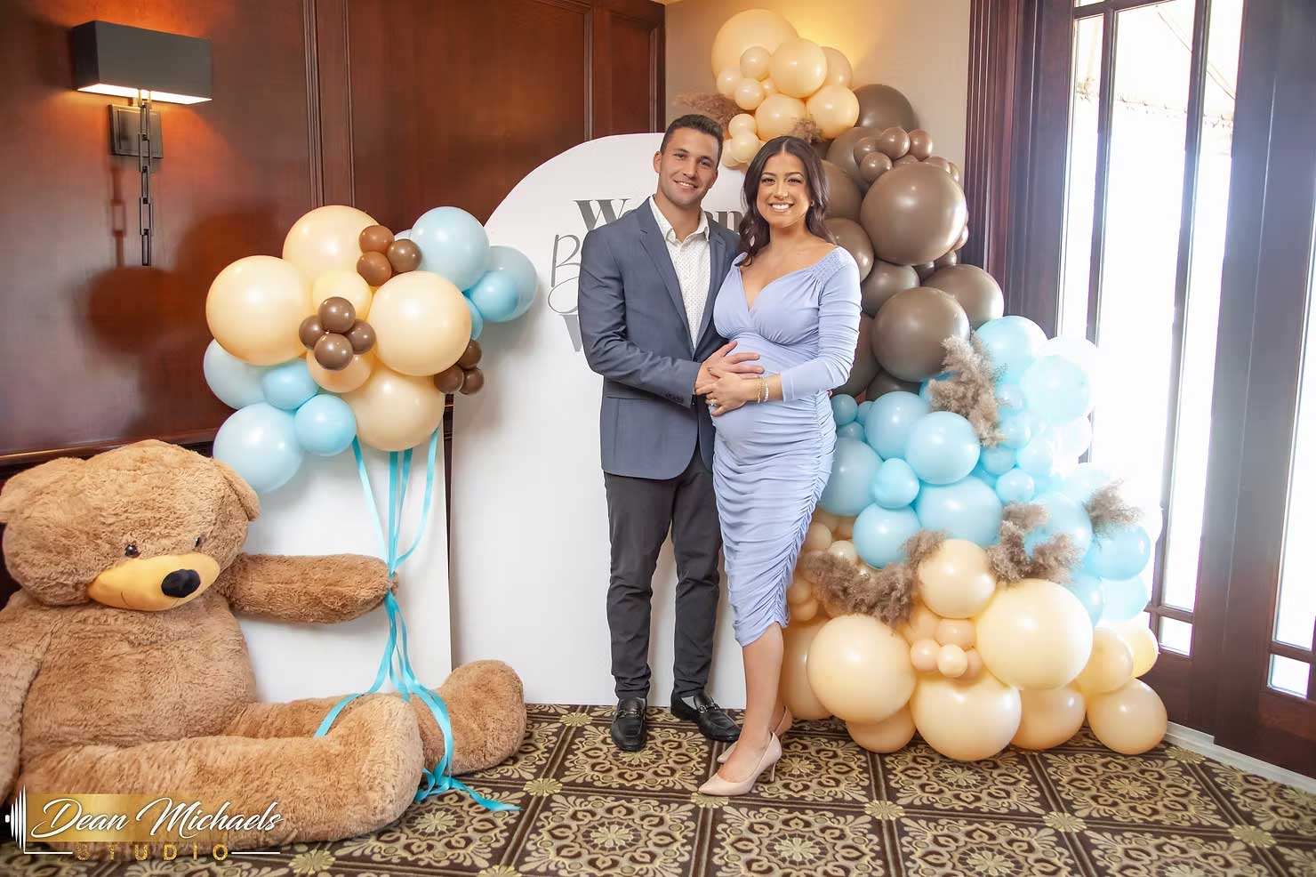brooklake-baby-shower-by-window-waterford-room A couple poses in front of a balloon display at an indoor NJ Bridal and Baby Showers event, with the woman holding her pregnant belly and a large teddy bear on the floor nearby.