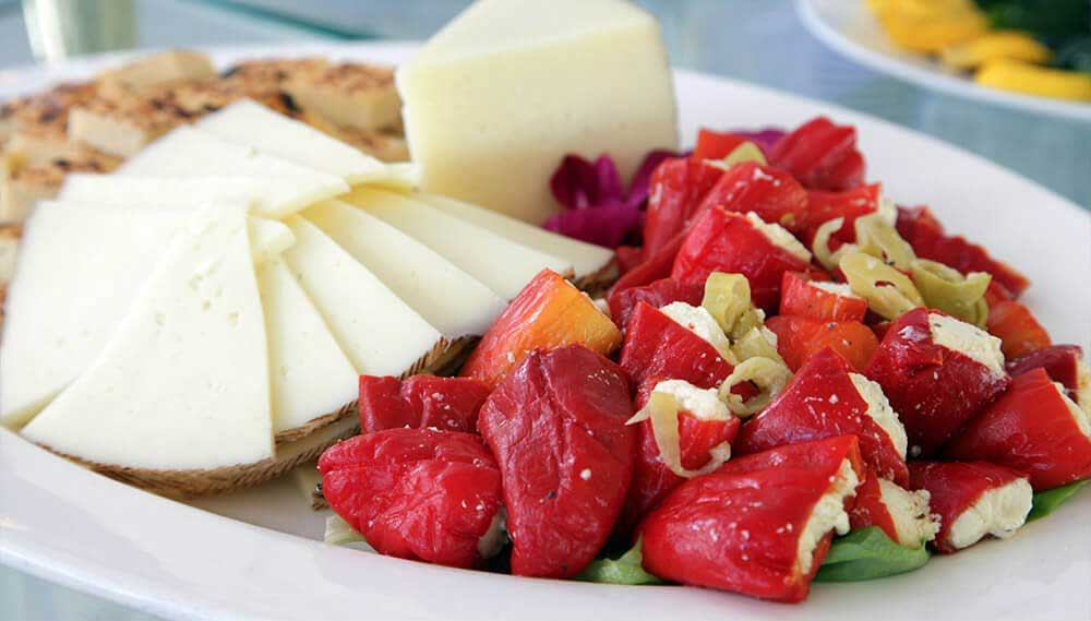 A plate with sliced white cheese, grilled bread, pickled vegetables, and a salad of chopped red and green peppers—perfect for Wedding & Event Cuisine.
