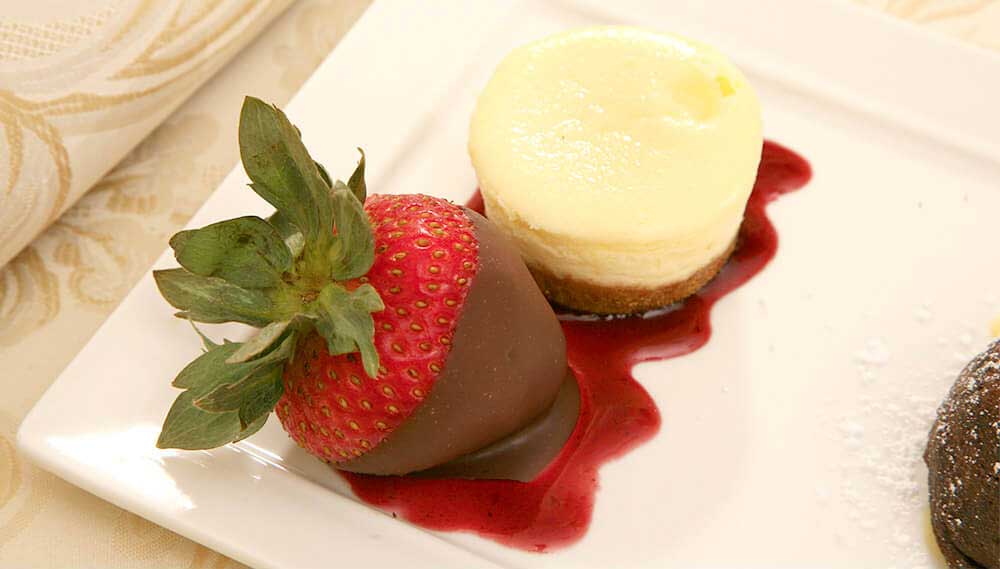 A chocolate-covered strawberry and a mini cheesecake sit on a white plate with berry sauce—an elegant touch of Wedding & Event Cuisine.