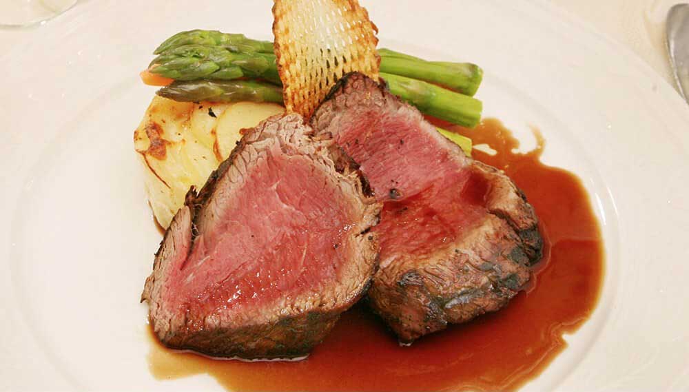 Two slices of medium-rare beef served with brown sauce, asparagus, potato gratin, and a crisp garnish on a white plate—a perfect choice for refined Wedding & Event Cuisine.