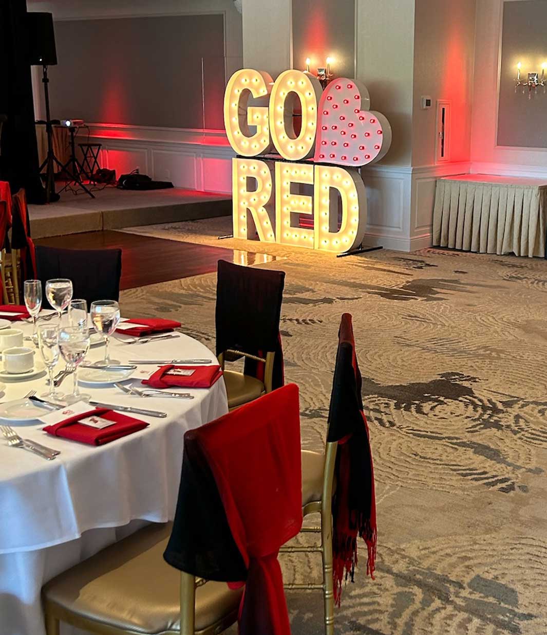 A banquet hall set for NJ corporate events features tables ready for guests; a large illuminated "GO RED" sign with a heart brightens the wall, while chairs are dressed in striking red and black covers.