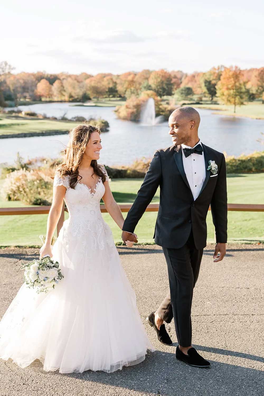 A bride and groom walk hand in hand outdoors on a sunny day, dressed in elegant wedding attire, with a lake and trees in the background—perfect for capturing timeless NJ Indian South Asian Weddings moments.