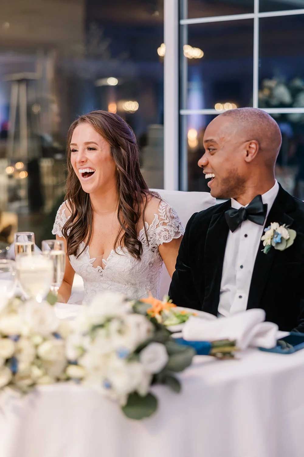 A bride and groom sit together at a table, smiling and laughing, with floral arrangements and glasses of champagne in front of them—capturing the joy of NJ Indian South Asian Weddings.