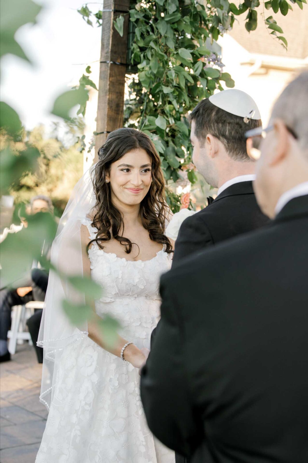 A bride and groom stand facing each other at an outdoor NJ Indian South Asian wedding ceremony, with the bride smiling and wearing a white dress and veil.