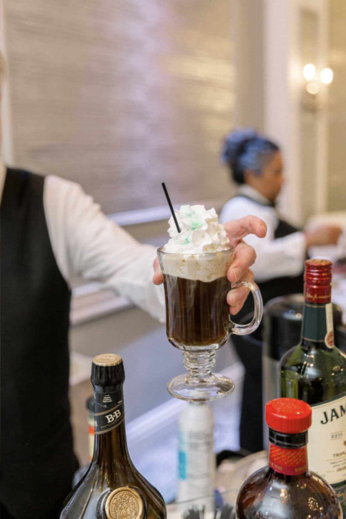 A server holds a glass mug of Irish coffee topped with whipped cream, with various bottles of liquor on the counter—perfect for warming up guests at NJ corporate events.