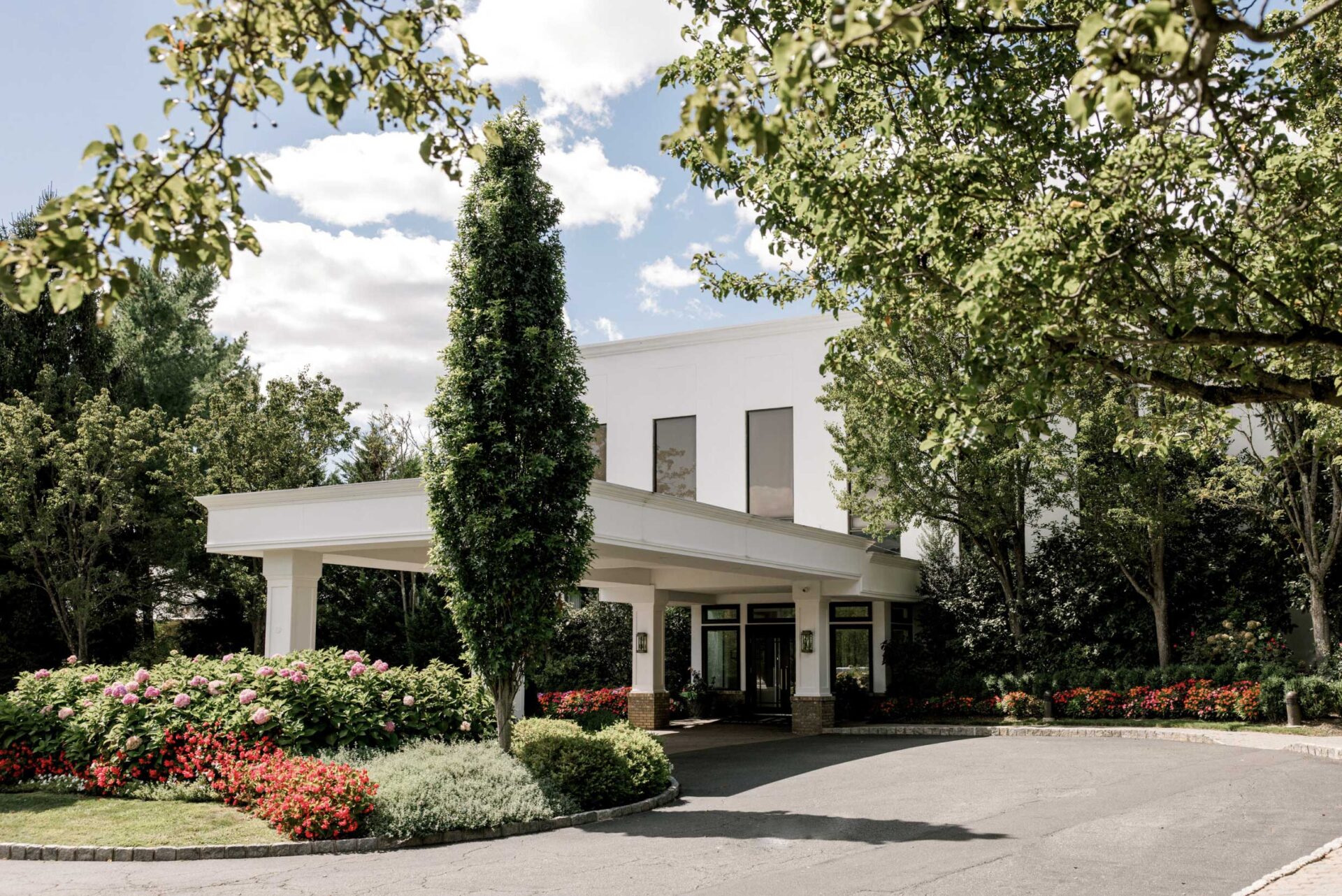A white building entrance with large windows, perfect for NJ Indian South Asian Weddings, is surrounded by manicured gardens, flower beds, and trees under a partly cloudy sky.