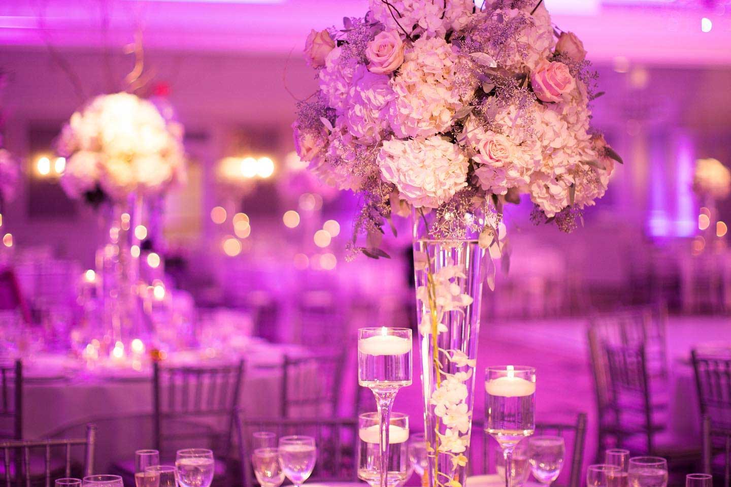 A tall centerpiece with pink and white flowers graces a table set with glassware and floating candles, under purple ambient lighting—perfect for elegant NJ Indian South Asian Weddings.