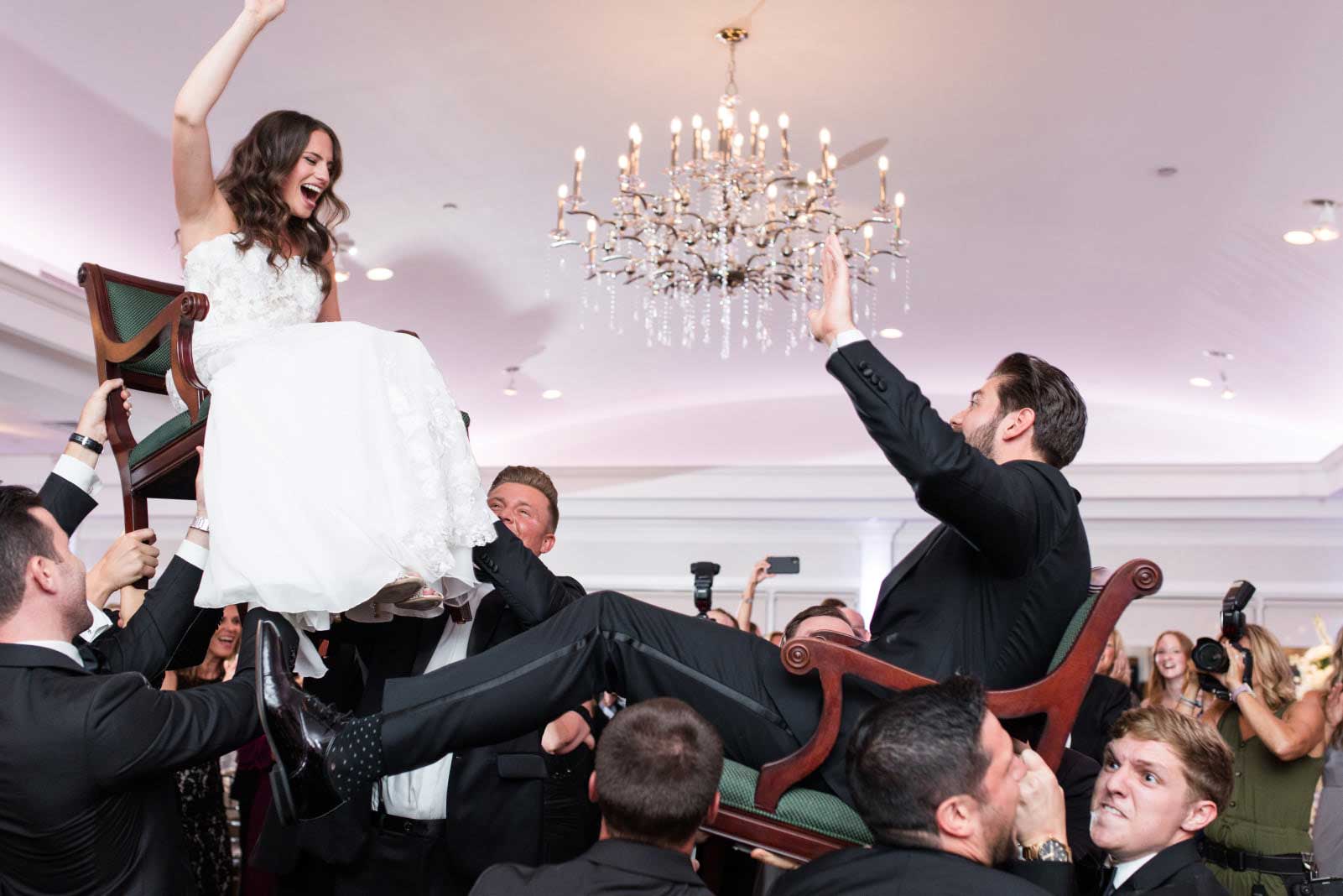 A bride and groom are joyfully lifted on chairs by guests during a vibrant NJ Kosher Weddings celebration in a brightly lit banquet hall.