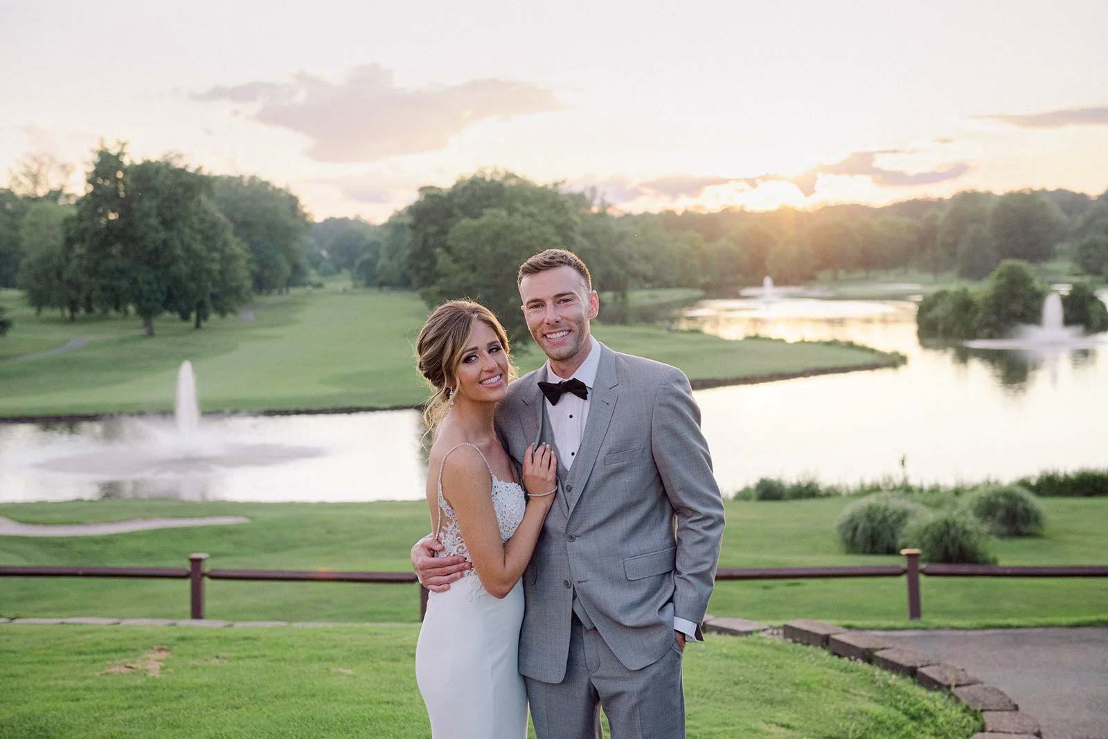 A couple poses in formal wedding attire outdoors at sunset, standing on grass with a pond, fountains, trees, and a golf course in the background—an ideal setting for unforgettable Wedding & Event Cuisine.