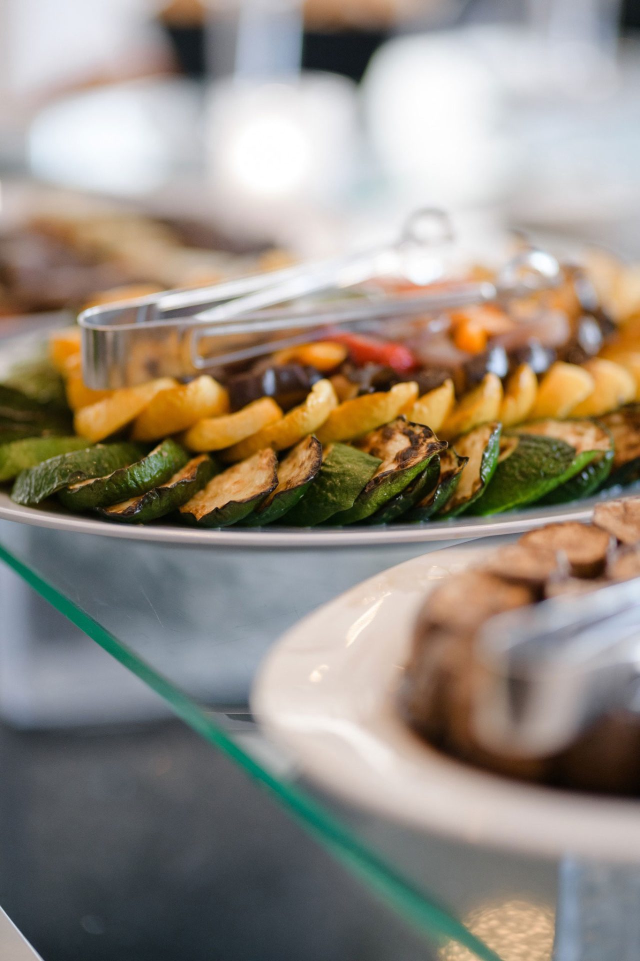 A platter of grilled vegetables, including zucchini and yellow squash, served with metal tongs on a buffet table—perfect for wedding & event cuisine.