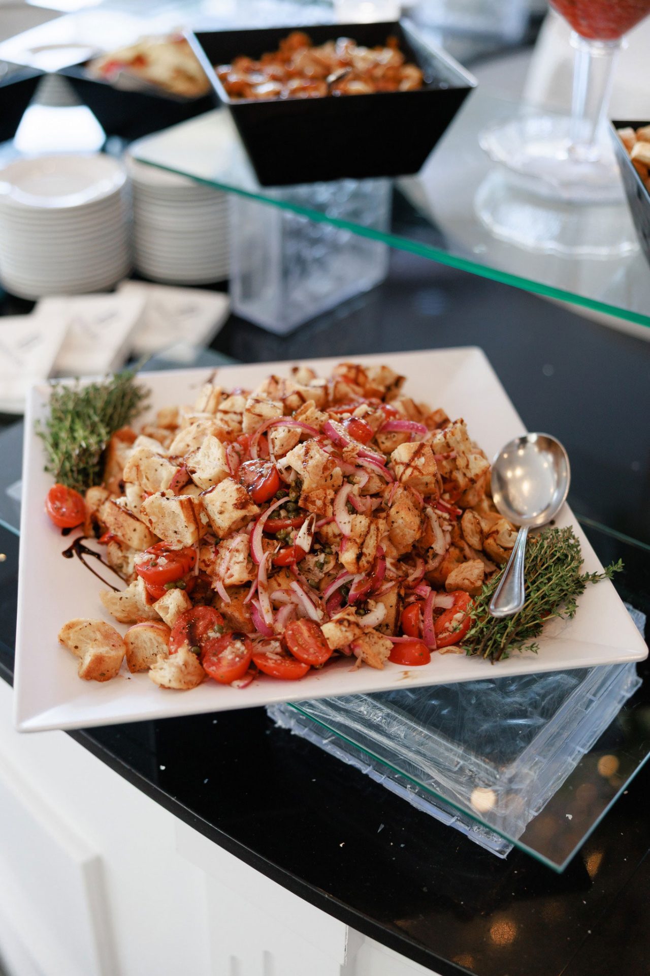 A square white plate with panzanella salad, featuring bread cubes, cherry tomatoes, red onions, herbs, and a serving spoon on the side—perfect for wedding & event cuisine.