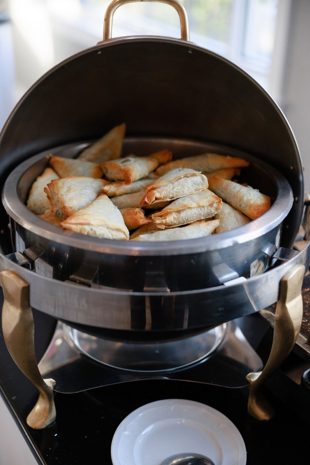 A metal chafing dish filled with golden-brown, triangular pastries sits on a buffet table next to an empty white plate, perfect for Wedding & Event Cuisine.